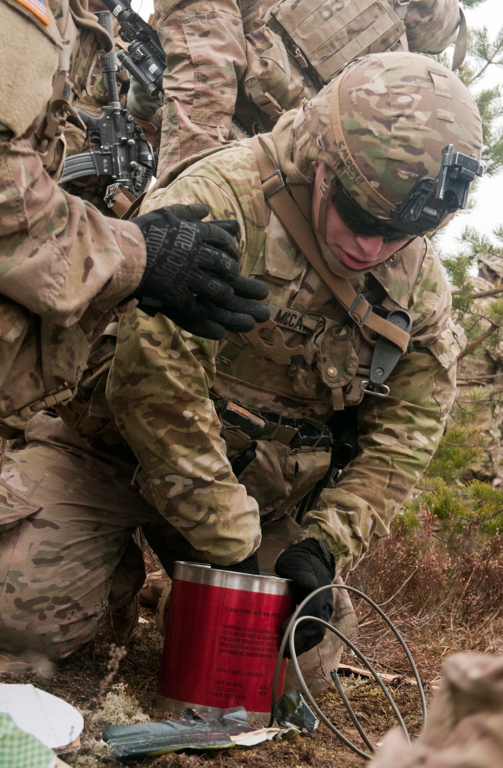A soldier in MultiCam/OCP camouflage uniform is crouching down and handling a red cylindrical demolitions or pyrotechnic device with attached wires/cables. The soldier, whose name tape partially reads 'MCCA-', is wearing a combat helmet with mounted equipment, Mechanix gloves, and full tactical gear. Additional soldiers are visible in the background, suggesting a training or operational exercise.