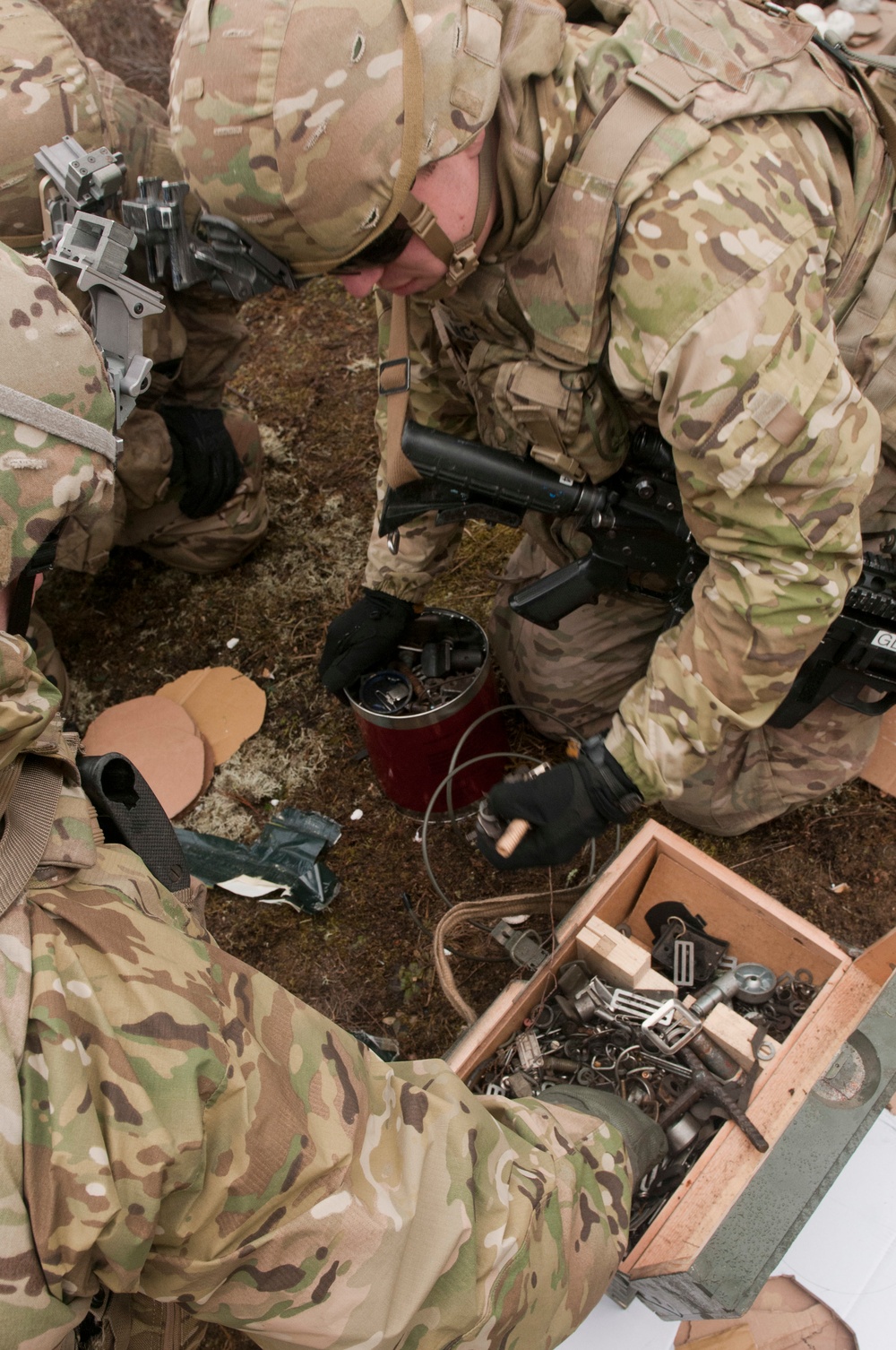Multiple soldiers in MultiCam/OCP camouflage uniforms are gathered around a ground-level work area, examining or assembling components from an open wooden crate containing various hardware and tools. The scene appears to be an outdoor training exercise involving equipment inspection or improvised device familiarization. A red container and loose wiring are also visible on the ground among the group.