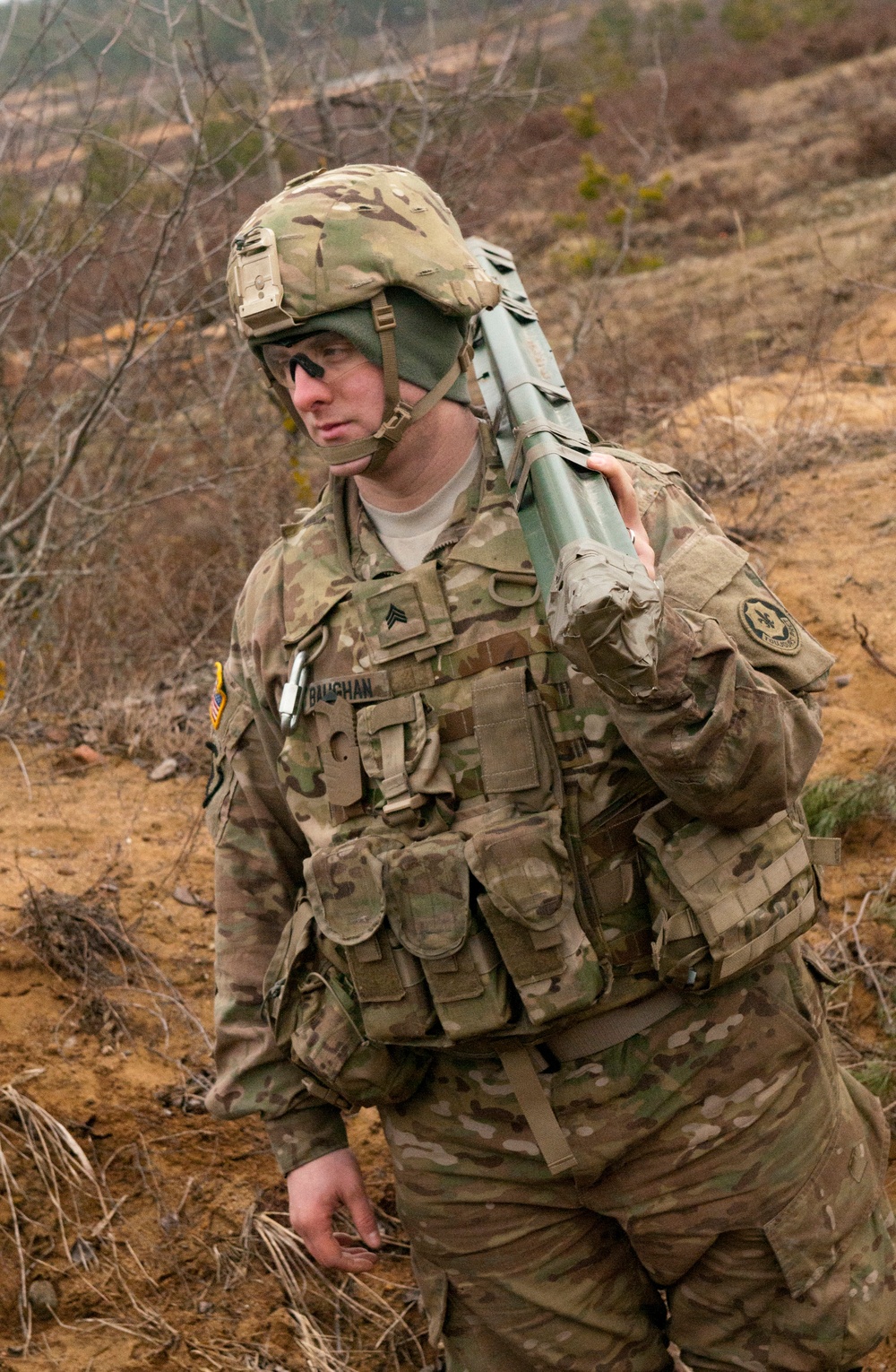 A U.S. Army soldier in full combat gear is moving through a sparse, arid outdoor training or operational environment. The soldier, identified by name tape as 'BAUGHAN' and wearing Sergeant rank insignia, is carrying what appears to be an AT4 anti-tank rocket launcher over his shoulder. He is wearing MultiCam (OCP) camouflage uniform with tactical vest, helmet with cover, and has face paint applied.