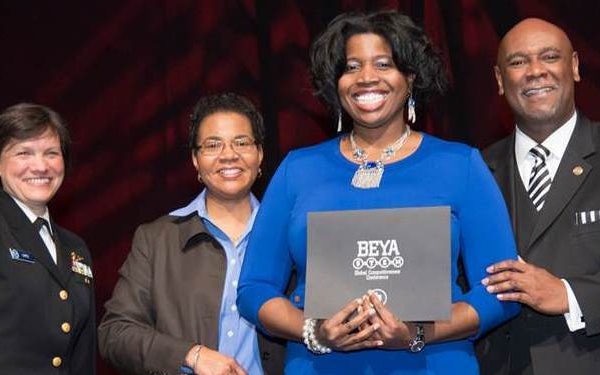 Four individuals are posing together at what appears to be a BEYA STEM Global Competitiveness Conference award ceremony. A woman in a blue dress is holding a BEYA STEM award folder/plaque while flanked by colleagues. The group appears to be on a stage with a red draped backdrop.