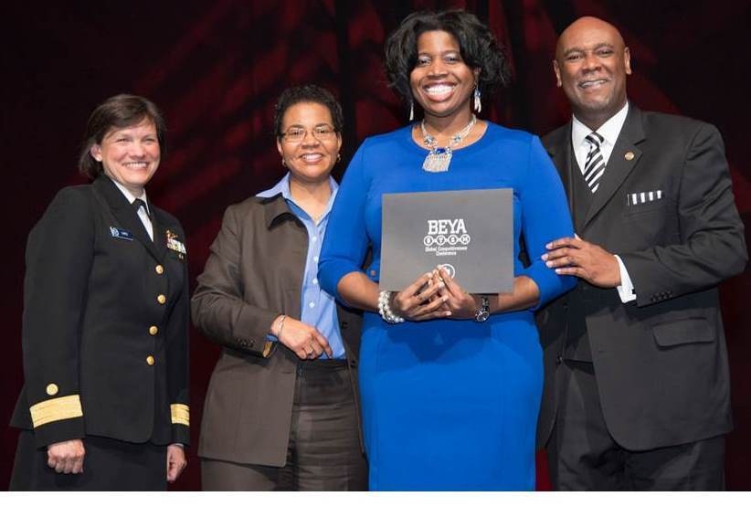 Four individuals are posing together at what appears to be a BEYA STEM Global Competitiveness Conference award ceremony. A woman in a blue dress is holding a BEYA STEM award folder/plaque while flanked by colleagues. The group appears to be on a stage with a red draped backdrop.