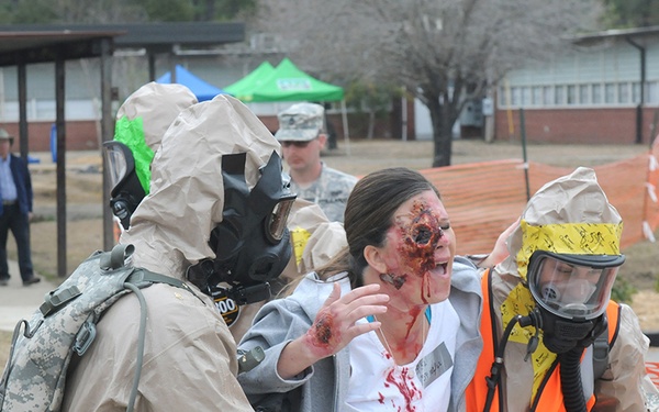 This image depicts a CBRN (Chemical, Biological, Radiological, Nuclear) emergency response training exercise or drill. Two responders in full HAZMAT suits and gas masks are assisting a simulated casualty wearing moulage (theatrical makeup) depicting facial and hand injuries with simulated blood. The setting appears to be a military or emergency management training facility with event tents and orange safety fencing visible in the background.