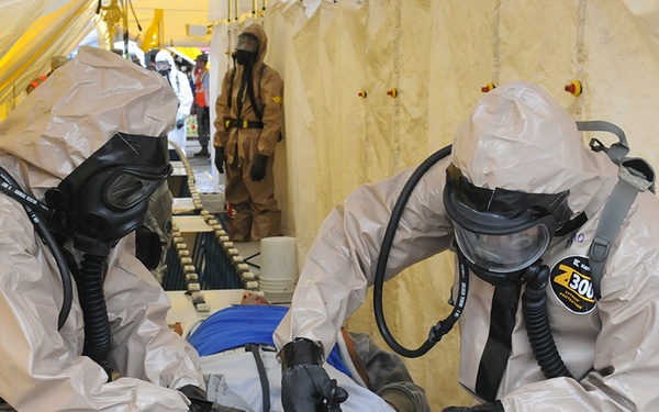 Two personnel in full CBRN (Chemical, Biological, Radiological, Nuclear) protective suits and gas masks are attending to a casualty lying on a table inside a yellow decontamination tent. The scene appears to be a training exercise or emergency response drill, with additional suited personnel visible in the background. The casualty's shoes and lower legs show white powdery residue, consistent with a simulated contamination scenario.