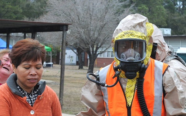 This image depicts what appears to be an emergency response drill or training exercise. A responder in full HAZMAT protective gear including a gas mask, tan suit, orange safety vest, and yellow tape seals is assisting civilian role-players wearing simulated injury makeup. The setting appears to be a military or government facility based on the brick buildings and uniformed personnel visible in the background.