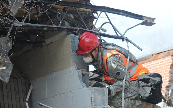 Two personnel in military camouflage uniforms and red safety helmets with orange high-visibility vests are conducting search and rescue operations in a heavily damaged building. The structure shows significant structural collapse with broken concrete block walls, twisted metal, and debris. The responders appear to be navigating through rubble, likely searching for survivors.