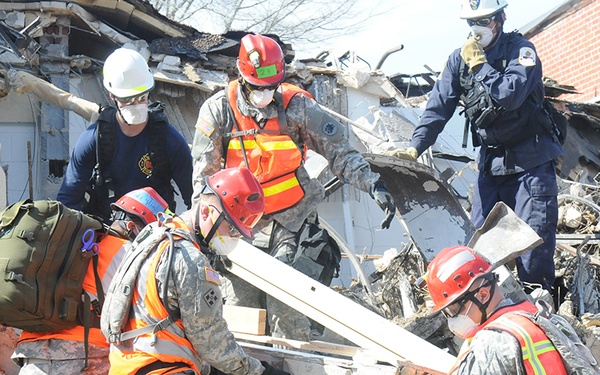 The image shows a search and rescue operation at a collapsed or heavily damaged brick building. Multiple responders in military camouflage uniforms and civilian emergency personnel are working together to clear debris. Personnel are wearing protective gear including hard hats, respirator masks, and high-visibility orange safety vests.