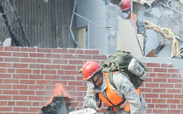 Military personnel in camouflage uniforms and safety gear are conducting urban search and rescue operations at a collapsed or heavily damaged brick building. At least three individuals are visible working through rubble, with one actively removing bricks from a hole in a wall. The scene depicts a disaster response or training exercise involving structural debris clearance.