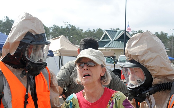 The image depicts what appears to be an emergency response drill or CBRN (Chemical, Biological, Radiological, Nuclear) training exercise. Two personnel wearing full hazmat suits with gas masks and high-visibility orange vests are assisting a civilian woman, likely acting as a simulated casualty or evacuee. The setting appears to be an outdoor staging area with tents, military personnel, and vehicles visible in the background.