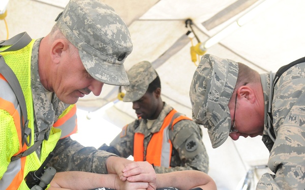 Military personnel in camouflage uniforms and high-visibility safety vests are attending to a civilian casualty lying on a stretcher or litter. The scene appears to be a medical training exercise or emergency response drill conducted under a tent structure. At least three uniformed individuals are actively working on the patient, who is wearing a dark civilian t-shirt.