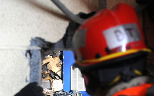 A person wearing military camouflage uniform and a red rescue helmet marked 'OT' is examining or working through a breach in a concrete block wall. The scene appears to be a search and rescue training exercise, with debris and damaged materials visible through the opening. An orange high-visibility vest is worn over the uniform, indicating a rescue or emergency response role.
