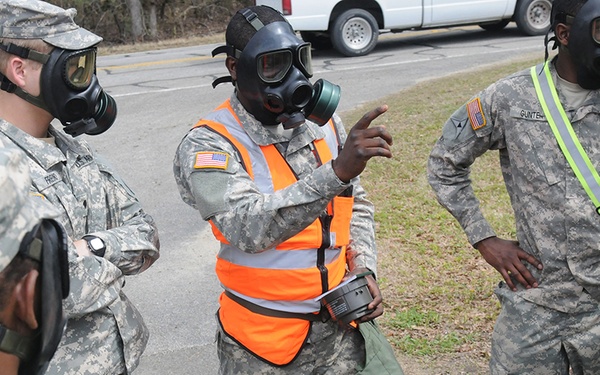 A group of U.S. Army soldiers wearing military combat uniforms and M40-series gas masks are gathered outdoors, apparently during a CBRN (Chemical, Biological, Radiological, Nuclear) training exercise. One soldier wearing an orange safety vest appears to be instructing or briefing the group, gesturing with one hand. The setting appears to be a roadside or training area with a white vehicle visible in the background.
