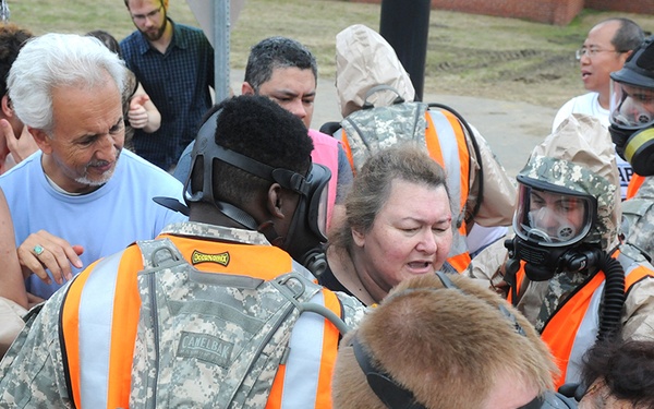 This image depicts what appears to be an emergency response drill or training exercise. Military personnel in camouflage uniforms with orange safety vests and gas masks are interacting with civilian role-players, some of whom are wearing simulated injury makeup. The scene includes hazmat-suited responders and multiple participants acting as casualties or bystanders.
