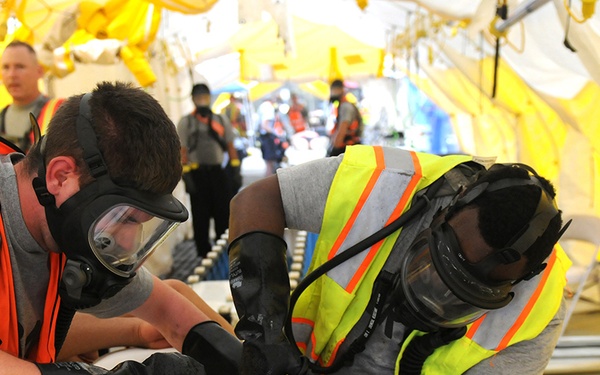 Two responders wearing full-face gas masks, safety vests, and black gloves are working on a patient lying on a stretcher or table, appearing to cut or remove clothing from the patient's legs. The scene takes place inside a large yellow tent structure, consistent with a mass casualty decontamination or emergency medical exercise. Additional personnel in safety vests are visible in the background, suggesting a coordinated multi-person drill or response operation.