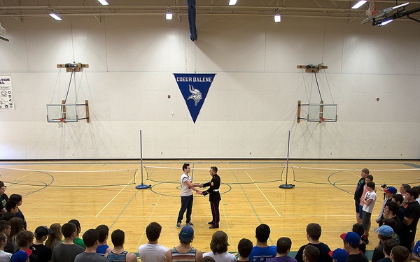 A large group of youth and young adults are gathered in a gymnasium at Coeur d'Alene school, forming a semicircle around two individuals who appear to be shaking hands at the center. The setting suggests a sports clinic, camp, or organized physical education event. A person in what appears to be a military uniform is one of the two individuals at the center of attention.