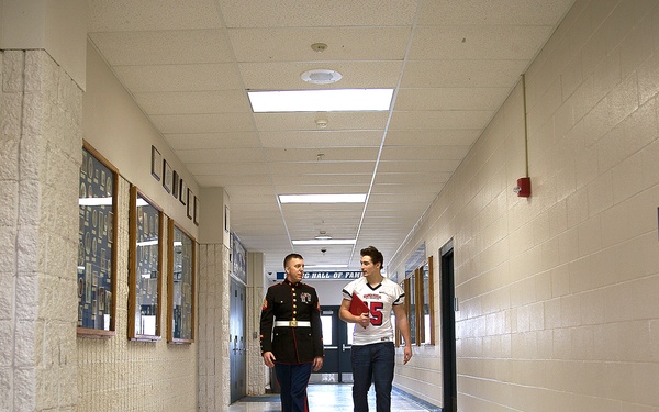 Two individuals are walking together down a school hallway. One person is wearing a U.S. Marine Corps dress uniform, while the other is a young man in a football jersey carrying what appears to be a red folder or binder. The hallway features trophy cases, framed photos, and a sign referencing a 'Hall of Fame.'