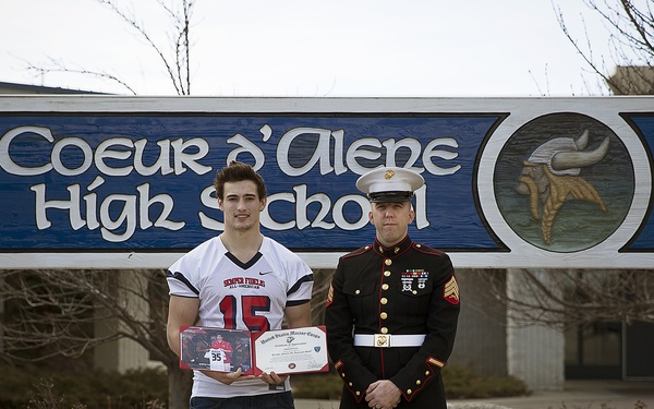 Two individuals stand in front of the Coeur d'Alene High School sign. A young man wearing a football jersey numbered 15 holds a framed award and certificate, standing alongside a United States Marine Corps servicemember in dress blues uniform. The setting appears to be a formal recognition or award presentation ceremony.