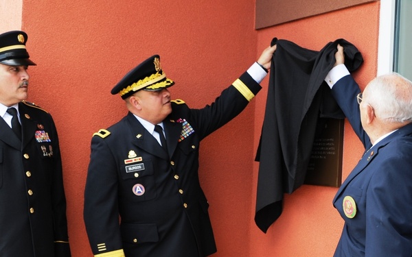 A formal military dedication ceremony is taking place at what appears to be a military installation. Two uniformed U.S. Army soldiers and two civilians are jointly unveiling a commemorative plaque mounted on a terracotta-colored wall. The plaque is being revealed by removing a black cloth covering.