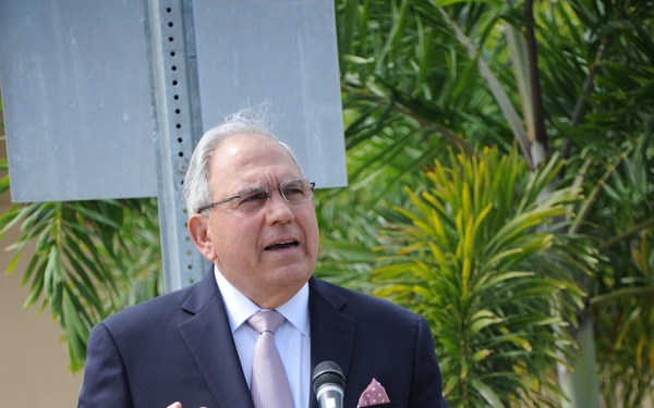 An older gentleman with gray hair and glasses is speaking at a podium during what appears to be an outdoor official event. The podium features a shield-shaped emblem displaying a red fire hydrant on a yellow background, suggesting a fire department or public safety organization. The setting includes palm trees and a building in the background, consistent with a warm-climate location.