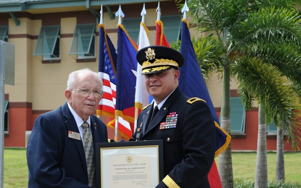 Two individuals are posing outdoors at what appears to be a U.S. Army military installation, jointly holding a framed Department of the Army Certificate of Appreciation. One person is an elderly civilian man in a blue suit, and the other is a U.S. Army officer in dress uniform wearing a general officer's cap and multiple service ribbons. The setting includes multiple flags, palm trees, and a military building in the background.