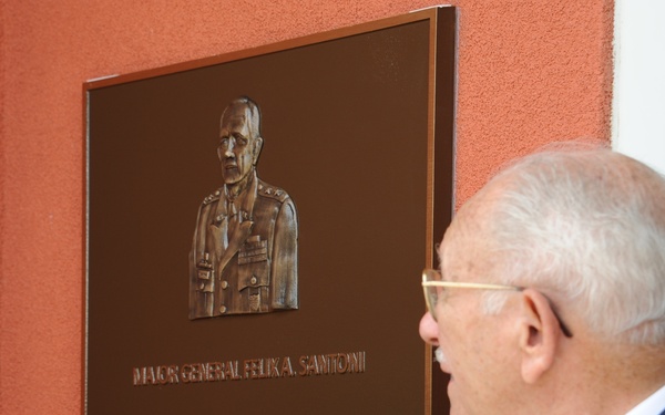 An elderly man in a dark suit is closely examining a bronze commemorative plaque mounted on a red/orange wall. The plaque honors Major General Felix A. Santoni and features a relief portrait of the general in military uniform along with an inscribed biographical text. The scene appears to be a dedication or unveiling ceremony.