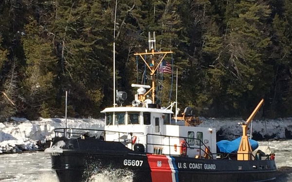 A U.S. Coast Guard vessel numbered 65607 is navigating through icy, partially frozen waters. The boat displays the distinctive USCG color scheme of black hull with red and blue diagonal stripe. The surrounding environment shows winter conditions with snow-covered shoreline and dense evergreen trees in the background.