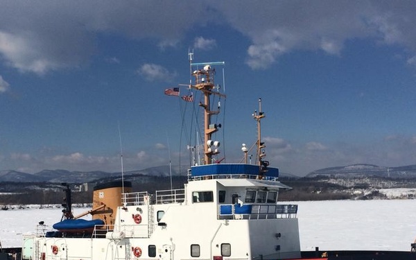 A U.S. Coast Guard icebreaker vessel numbered 108 is navigating through heavily iced waters, surrounded by broken ice chunks. The ship displays the distinctive USCG diagonal racing stripe in red, white, and blue on its black hull. Snow-covered mountains and a partly cloudy blue sky are visible in the background.