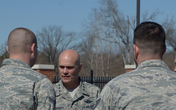 A group of five U.S. Air Force personnel in ABU (Airman Battle Uniform) camouflage uniforms are gathered in an outdoor setting, appearing to be in a briefing or discussion. The central figure, facing the camera, displays general officer rank insignia (two stars) and a name tape partially reading 'RHART' with 'US AIR FORCE' visible. The setting appears to be a military installation with a fence and bare trees in the background.
