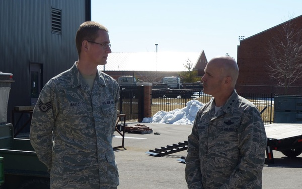 Two U.S. Air Force personnel in Airman Battle Uniforms (ABU) are engaged in conversation outdoors on a paved surface at what appears to be a military installation. The individual on the left has a name tape reading 'BAATZ' and wears Staff Sergeant chevrons, while the individual on the right appears to wear general officer rank insignia (two stars visible). Snow piles are visible in the background, suggesting a winter or early spring setting.
