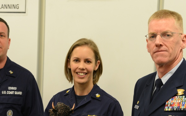 Three U.S. Coast Guard personnel are posing for a formal photo in what appears to be a planning room or briefing area. The central figure, a female service member, is holding a trophy or award featuring what appears to be a bronze eagle or bison figurine on a black base. The officer on the right is in a formal dress uniform with an extensive array of service ribbons and medals.