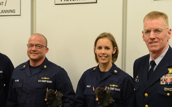 Four U.S. Coast Guard personnel pose for a formal photo, with two members holding eagle-topped trophies or awards. The setting appears to be an indoor facility with whiteboards visible in the background displaying text related to 'Risk Assessment/Planning' and 'Prevention'. The rightmost individual wears a formal dress uniform with extensive medal ribbons, suggesting senior rank.