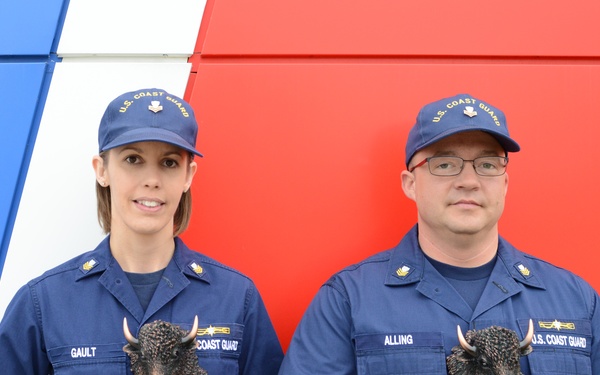 Two U.S. Coast Guard personnel in blue uniforms are posing with trophy awards in front of a red, white, and blue painted wall. Their name tapes read 'GAULT' (female, left) and 'ALLING' (male, right), and both are holding identical bronze bison head trophies on black bases. The plaques on the trophies appear to indicate 'Volunteer Person of the Year' awards.