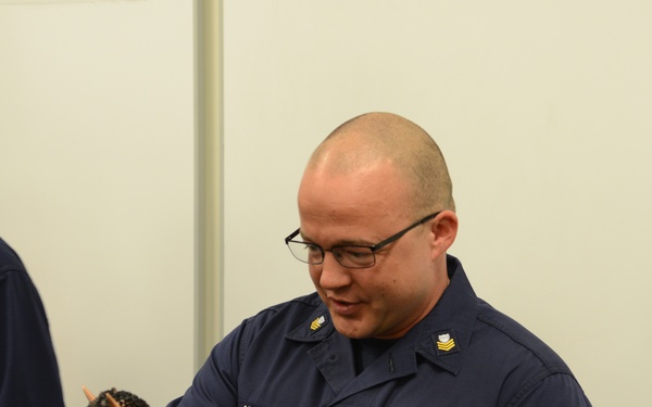A U.S. Coast Guard petty officer named Alling is holding and examining a bronze animal figurine trophy or award mounted on a dark base. He is wearing a navy blue Coast Guard uniform with visible rank insignia, a name tape, branch tape, a qualification badge, and a medal with a green and orange ribbon. The setting appears to be an indoor facility, likely during an awards or recognition ceremony.