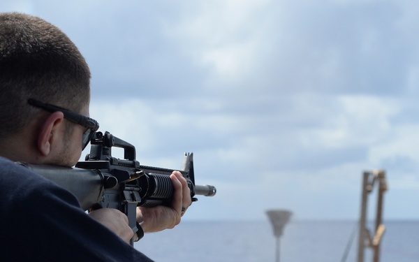 A person in a dark navy sweatshirt is aiming an M16-style rifle on what appears to be the deck of a naval vessel at sea. A paper shooting target mounted on a wooden stand is visible downrange. This appears to be a marksmanship qualification or training exercise conducted aboard a ship.