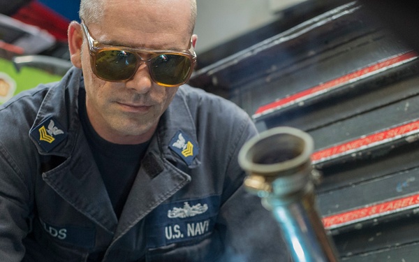 A U.S. Navy petty officer is performing torch welding or brazing work in what appears to be a workshop or maintenance facility aboard a ship or naval installation. He is wearing protective tinted safety glasses and a dark blue Navy coverall uniform with rank insignia and a name tag. The image captures him focused on his work with a lit torch, with visible smoke and a metal component being worked on.