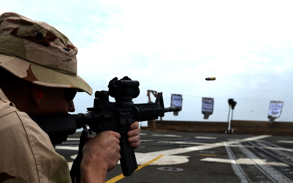 A person in desert camouflage uniform and boonie hat is firing a rifle in the prone position on what appears to be the deck of a naval vessel. A spent brass casing is visibly ejecting from the weapon mid-air. Shooting targets are set up downrange along the ship's deck.
