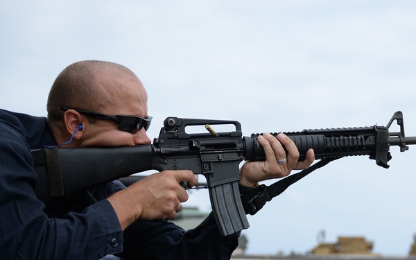 A person in dark blue uniform is lying in a prone shooting position on a blue mat, aiming an M16-style rifle. The setting appears to be an outdoor firing range, likely on a military vessel or installation based on the surface and background structures. A second person wearing sunglasses is partially visible in the background.