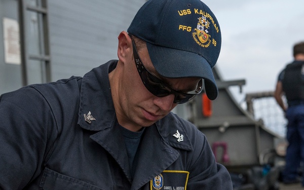 A U.S. Navy sailor assigned to USS Kauffman (FFG-59) is seated on the ship's deck, handling and inspecting what appears to be a pistol. He is wearing Navy working uniform (NWU) with a name tag reading 'Daniel L. Anderson' and a USS Kauffman ball cap. Additional personnel are visible in the background on the ship's deck.