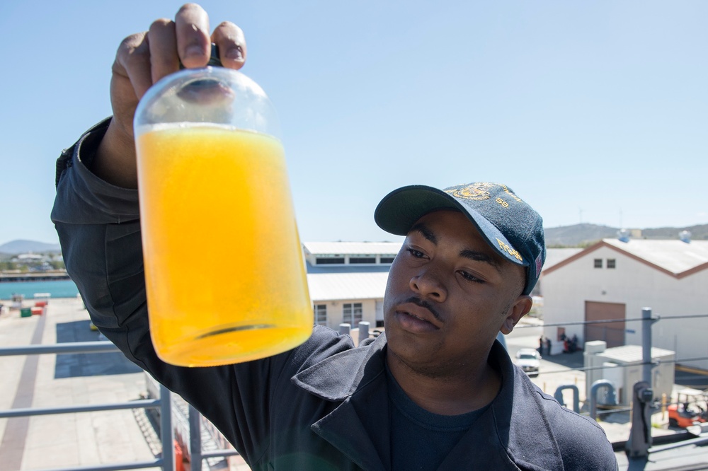 A person wearing a dark uniform and a navy-style cap holds up a glass jar containing a bright orange-yellow liquid, examining it closely. The setting appears to be a military or industrial facility, likely a naval base, with buildings and equipment visible in the background. The individual appears to be conducting a quality control or inspection check of the liquid sample.