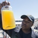 A person wearing a dark uniform and a navy-style cap holds up a glass jar containing a bright orange-yellow liquid, examining it closely. The setting appears to be a military or industrial facility, likely a naval base, with buildings and equipment visible in the background. The individual appears to be conducting a quality control or inspection check of the liquid sample.