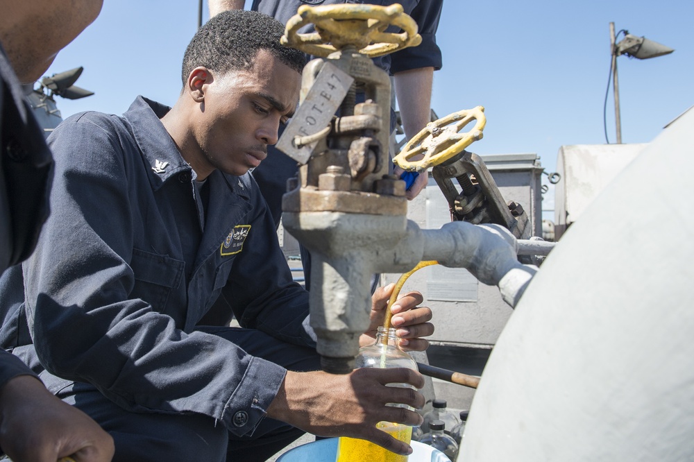 A U.S. Navy sailor in dark blue coveralls is collecting a fluid sample from a shipboard valve/pipe system into a clear glass jar. The sample appears to be a yellow-amber liquid, likely fuel or hydraulic fluid, being drawn through a hose connected to an industrial valve assembly. The setting appears to be the exterior deck of a naval vessel under clear blue skies.