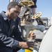 A U.S. Navy sailor in dark blue coveralls is collecting a fluid sample from a shipboard valve/pipe system into a clear glass jar. The sample appears to be a yellow-amber liquid, likely fuel or hydraulic fluid, being drawn through a hose connected to an industrial valve assembly. The setting appears to be the exterior deck of a naval vessel under clear blue skies.