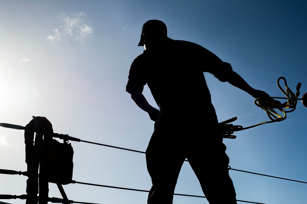 A silhouetted figure, likely a sailor or dockworker, is captured from a low angle against a bright blue sky while handling a rope or line. The strong backlit composition creates a dramatic silhouette effect, obscuring fine details but emphasizing the physical action and working environment. Railing lines, rigging equipment, and what appears to be a life preserver are visible in the background, suggesting a maritime vessel setting.