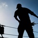 A silhouetted figure, likely a sailor or dockworker, is captured from a low angle against a bright blue sky while handling a rope or line. The strong backlit composition creates a dramatic silhouette effect, obscuring fine details but emphasizing the physical action and working environment. Railing lines, rigging equipment, and what appears to be a life preserver are visible in the background, suggesting a maritime vessel setting.