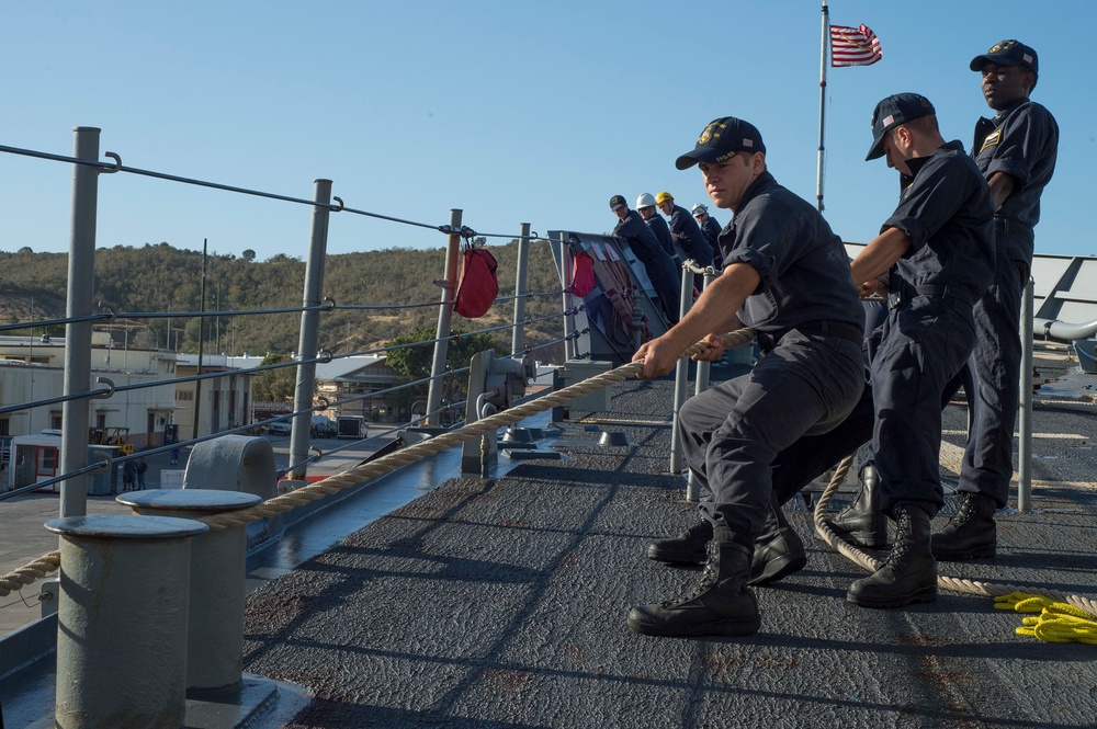 A group of uniformed naval personnel are on the deck of a military vessel, pulling a thick mooring rope in a coordinated effort. The scene appears to be a docking or undocking operation at a naval facility, with a US flag visible on a flagpole in the background. The setting appears to be a port or naval base with buildings and hills visible in the background.