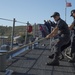 A group of uniformed naval personnel are on the deck of a military vessel, pulling a thick mooring rope in a coordinated effort. The scene appears to be a docking or undocking operation at a naval facility, with a US flag visible on a flagpole in the background. The setting appears to be a port or naval base with buildings and hills visible in the background.