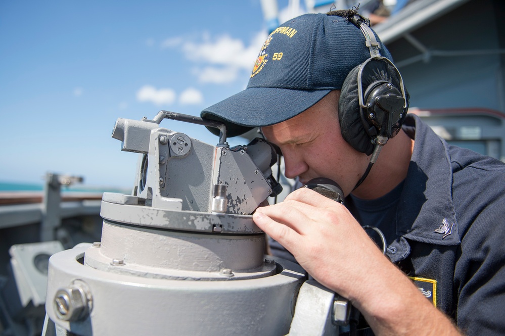 A U.S. Navy sailor is using a gyrocompass or bearing circle on the bridge wing of a naval vessel. The sailor is wearing a navy blue cap with '59' visible and communication headphones, suggesting active watch duties. The setting is an open deck of a warship under clear blue skies.