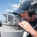 A U.S. Navy sailor is using a gyrocompass or bearing circle on the bridge wing of a naval vessel. The sailor is wearing a navy blue cap with '59' visible and communication headphones, suggesting active watch duties. The setting is an open deck of a warship under clear blue skies.