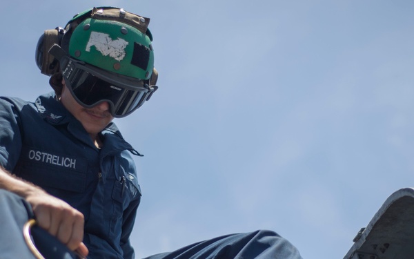 A U.S. Navy sailor identified by name tape as 'OSTRELICH' is performing maintenance work on what appears to be aircraft or shipboard equipment. The sailor is wearing a green flight deck helmet with goggles and a blue Navy utility uniform while handling a tool or lubrication device. The image is taken from a low angle against a partly cloudy sky.