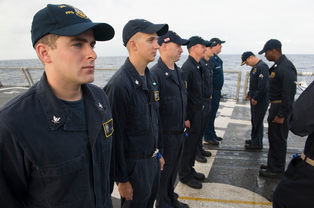 A group of U.S. Navy sailors in dark blue utility uniforms (NWU Type III or similar) are standing in formation on the deck of a naval vessel, likely during an inspection or muster. The ship appears to be underway at sea, visible from the ocean horizon in the background. One sailor on the far right appears to be holding a clipboard, suggesting an administrative or inspection activity.