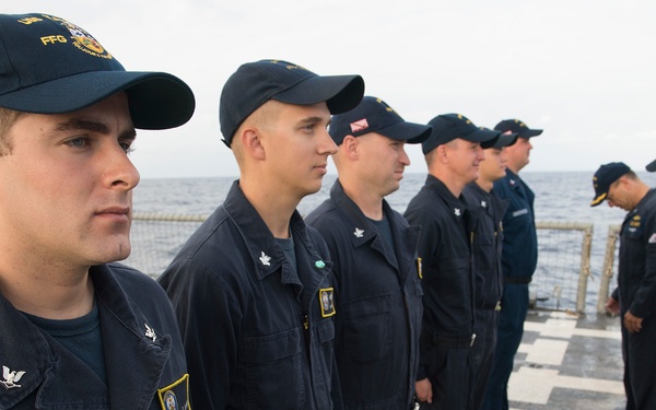 A group of U.S. Navy sailors in dark blue utility uniforms (NWU Type III or similar) are standing in formation on the deck of a naval vessel, likely during an inspection or muster. The ship appears to be underway at sea, visible from the ocean horizon in the background. One sailor on the far right appears to be holding a clipboard, suggesting an administrative or inspection activity.